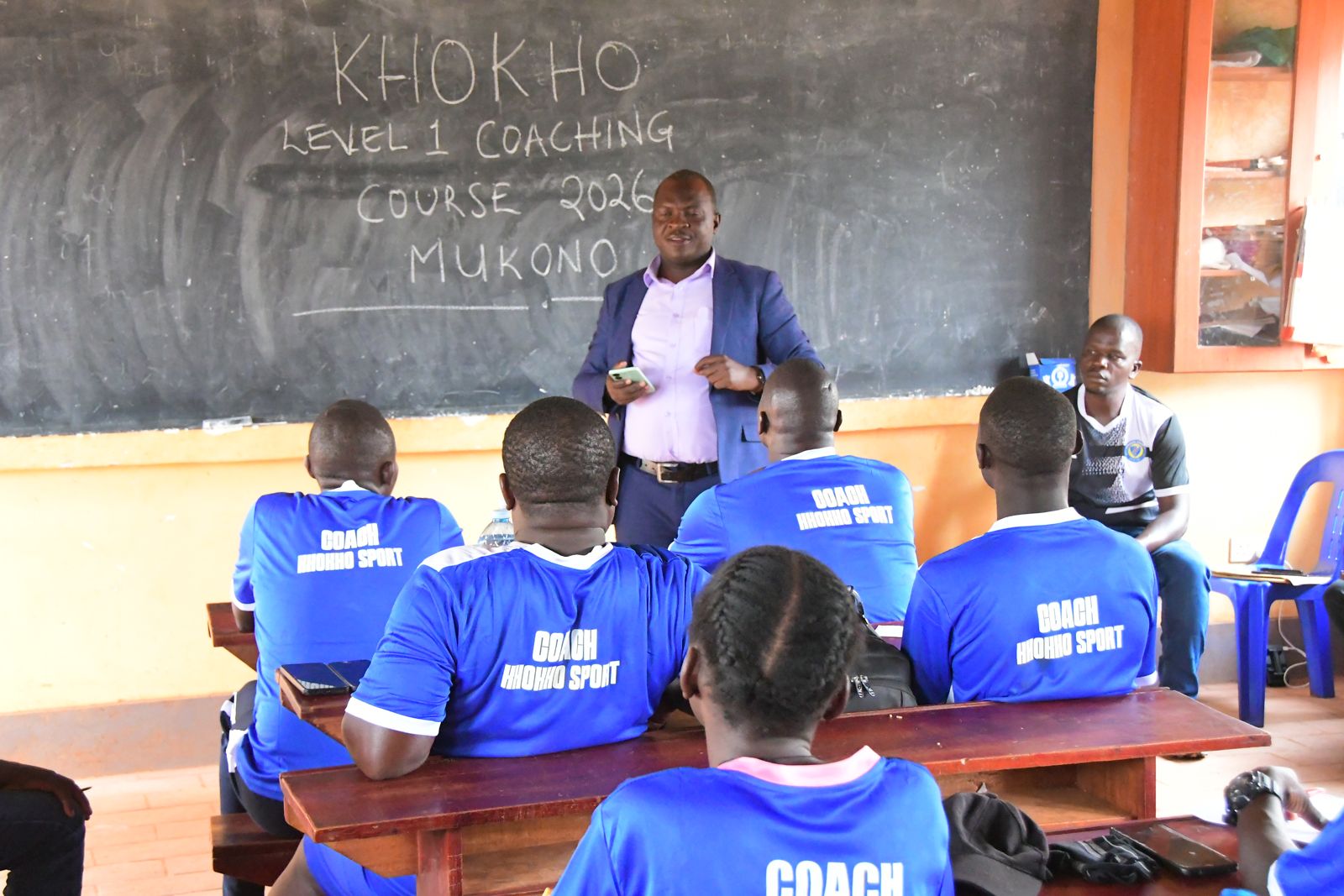 A trainer addresses participants in front of a blackboard reading Khokho Level 1 Coaching Course 2026 Mukono